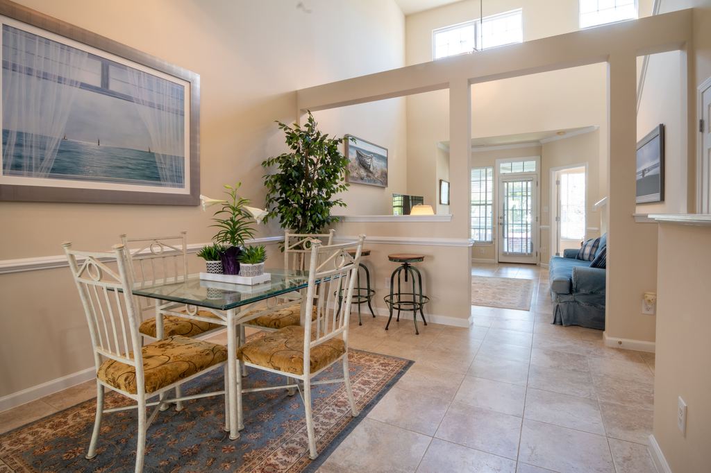 Coastal dining area with glass table, white chairs, beach artwork, indoor plants, and soft blue accents in an open-plan home.