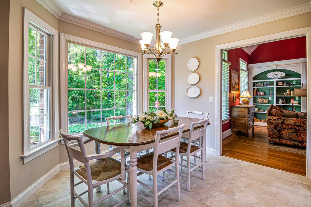 Bright traditional dining room with large windows, round wooden table, white chairs, chandelier, and an adjoining red living room.