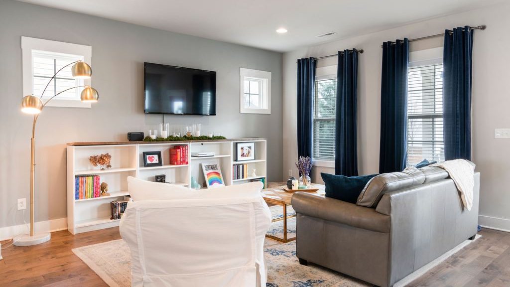 Rental living room with layered lighting, dark curtains, wall-mounted TV, area rug, storage console, and mixed seating.