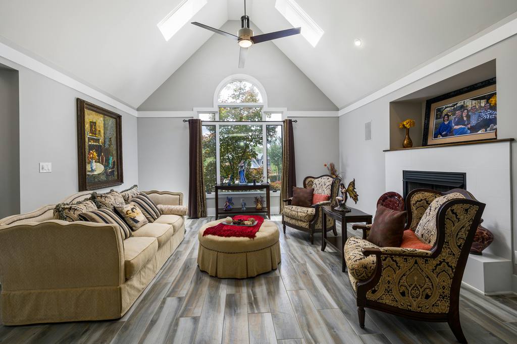 Traditional living room with vaulted ceiling, patterned armchairs, cream sofa, round ottoman, fireplace, large window, and classic upholstered seating.
