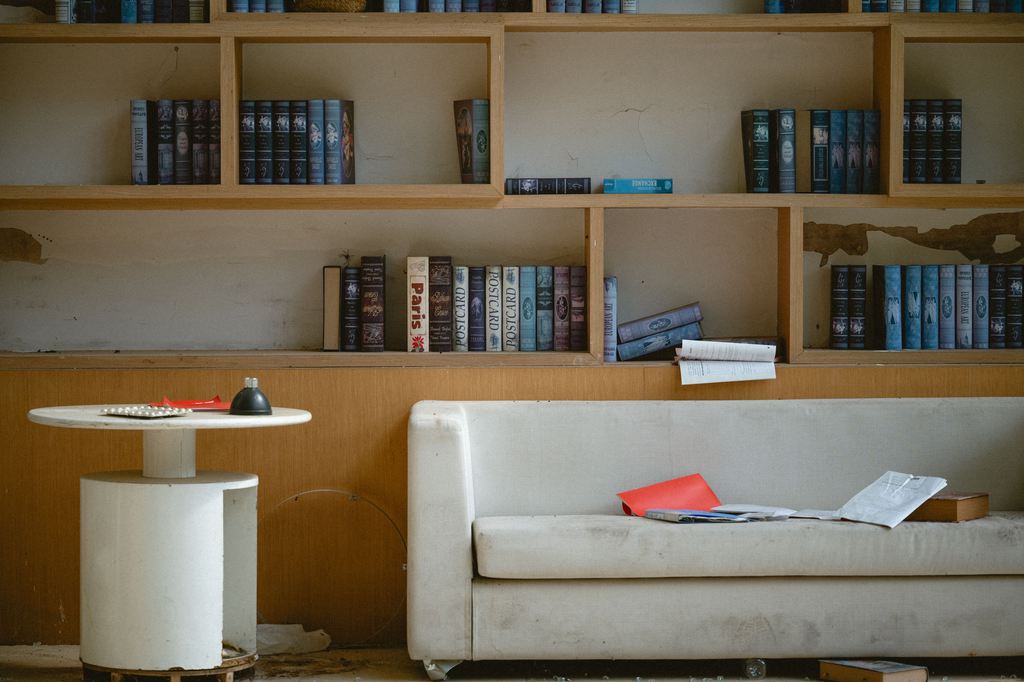 Worn white sofa beneath an unfinished wood bookcase with scattered books and papers in a neglected living room.