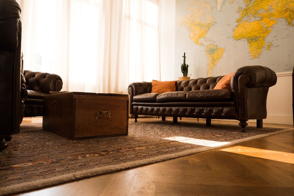 Dark brown leather Chesterfield sofa with orange pillows, wooden trunk coffee table, patterned rug, and world map wall art in a warm living room.
