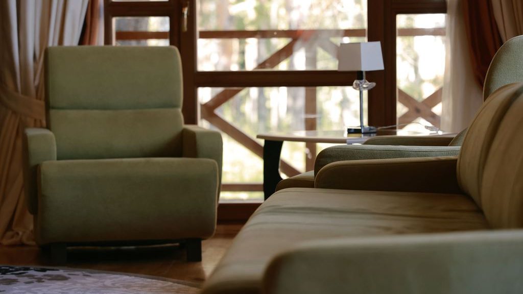 Cozy sitting area with green upholstered armchairs, a small side table, and soft daylight through tall wood-framed windows.