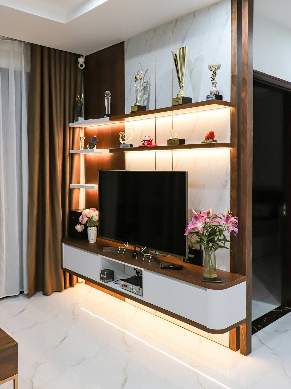 Modern TV stand with open shelving, warm lighting, trophy decor, and floral accents in a polished living room setup.