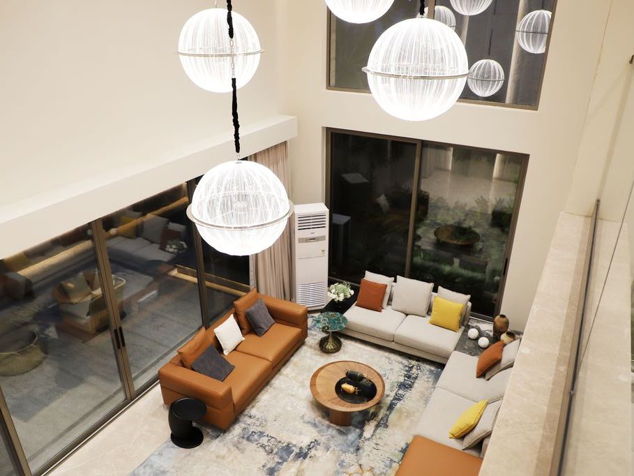 Double-height living room with tan and cream sofas, a round wood coffee table, oversized globe pendant lights, and floor-to-ceiling glass doors.