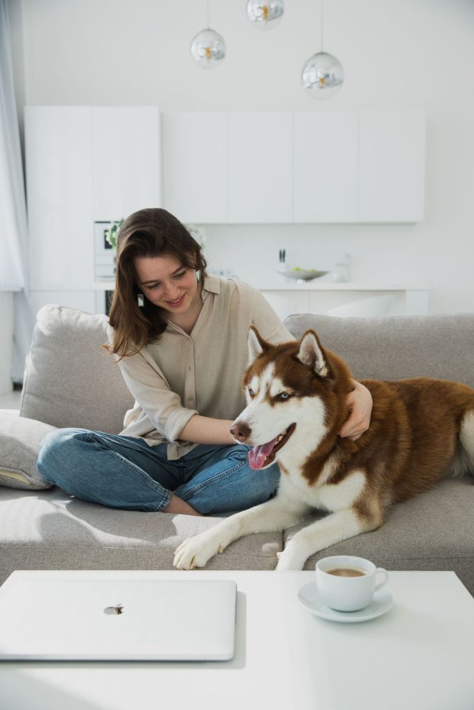 Woman sitting with a large husky on a light gray fabric sofa beside a coffee table.