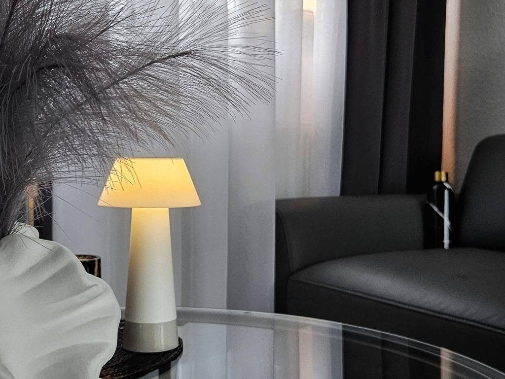 Modern minimalist lounge with black sofa, glass table, soft table lamp, layered curtains, and sculptural white vase.