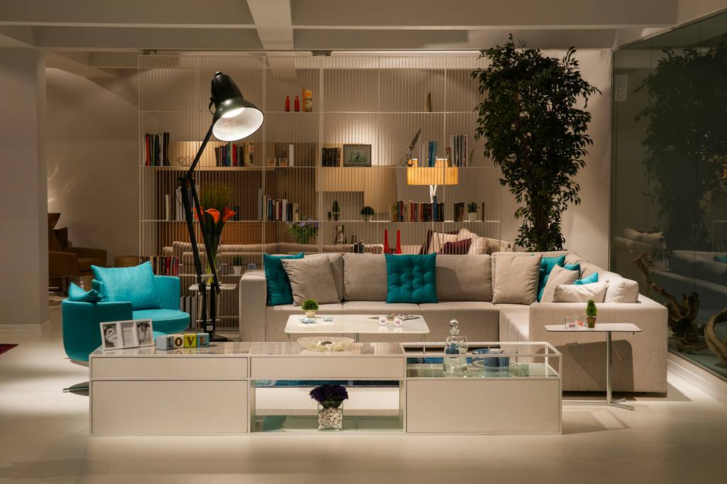 Modern living room with a beige sectional sofa, turquoise pillows and accent chair, white coffee tables, floor lamp, shelving, and indoor greenery.