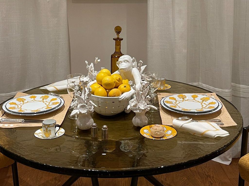 Round dark glass dining table set with yellow-patterned place settings, a white fruit bowl filled with lemons, and decorative white ceramic accents.