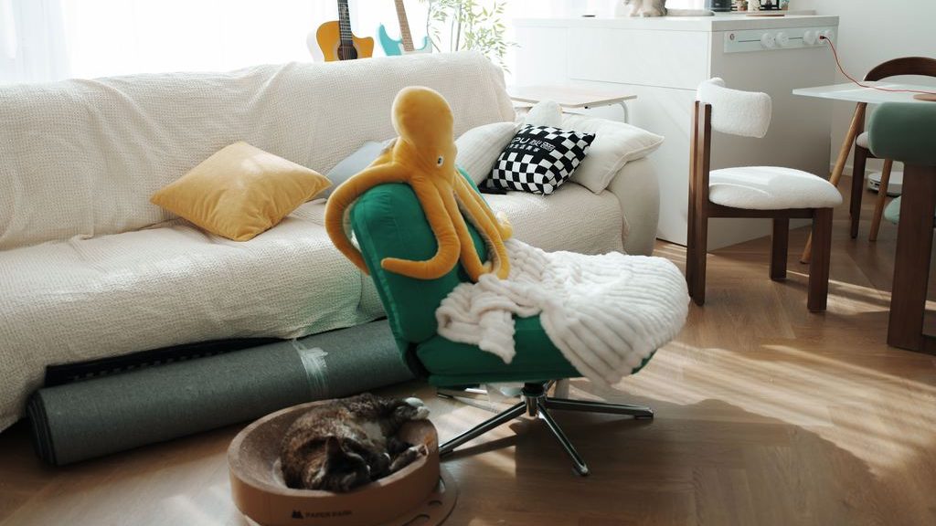 Green swivel office chair beside a white sofa in a bright apartment living room, with a cat bed, dining table, and colorful cushions.