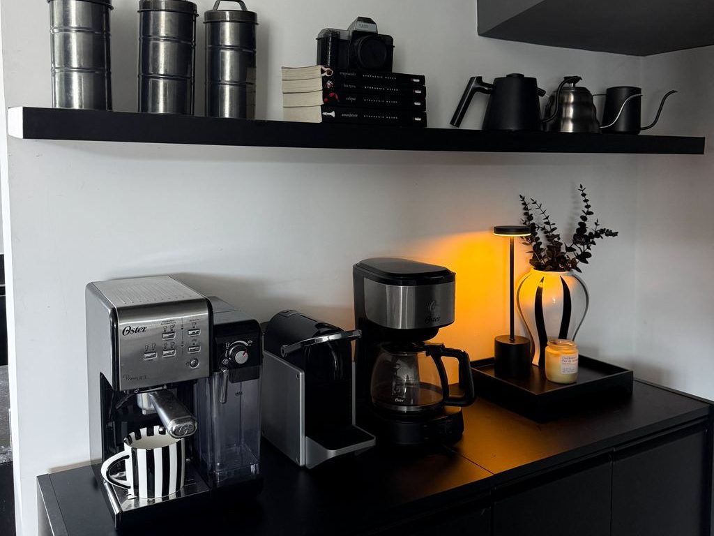 Black home coffee bar with espresso machine, drip coffee maker, floating shelf storage, books, teapots, and warm accent lighting.