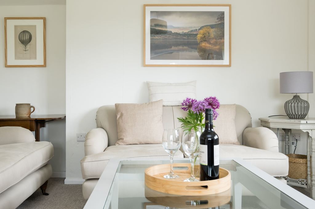 Wooden coffee table tray with wine, glasses, and flowers on a glass table in a cozy neutral living room.