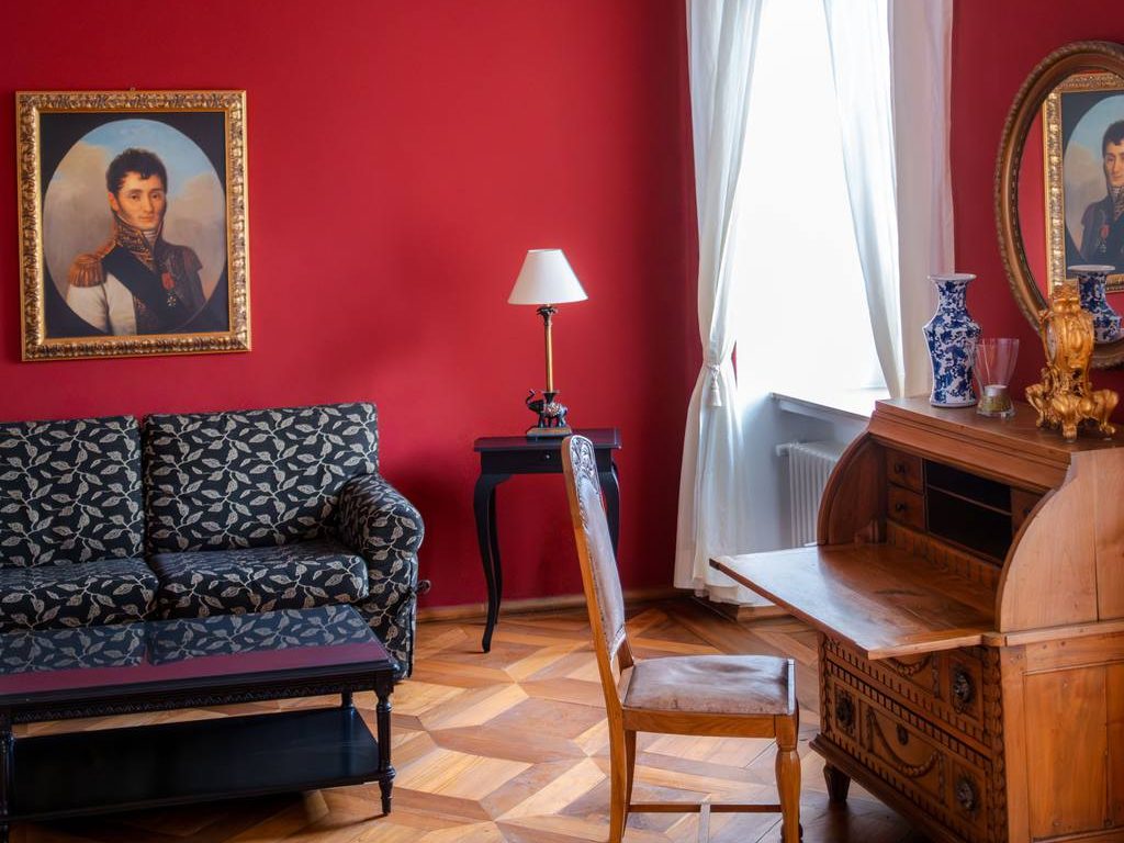 Traditional interior design room with red walls, patterned sofa, antique wooden desk, framed portraits, sheer curtains, and classic parquet flooring.