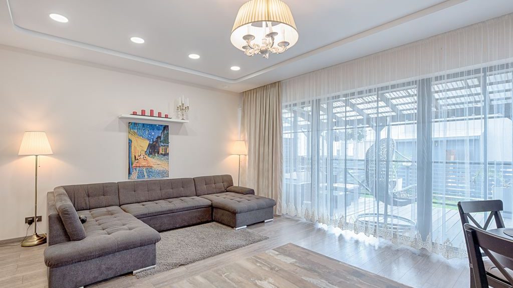 Gray U-shaped sectional sofa in a bright modern living room with floor lamps, sheer curtains, a large window, and light wood flooring.
