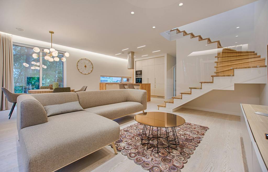 Modern open-plan living room with gray sectional sofa, round coffee table, patterned rug, white staircase, and warm wood accents.