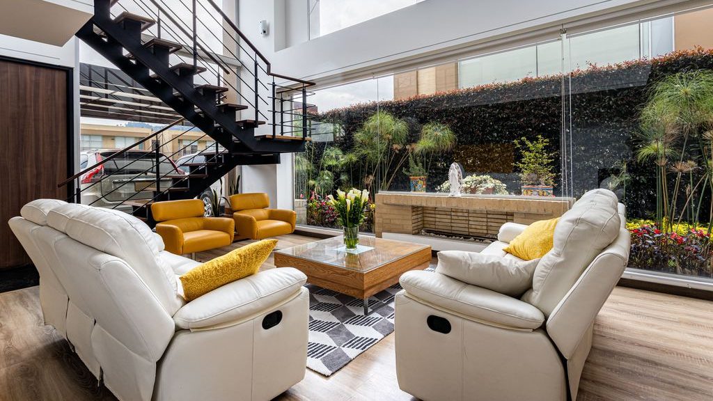 Bright double-height living room with white leather recliners, yellow armchairs, a wood coffee table, and floor-to-ceiling windows facing an indoor garden.