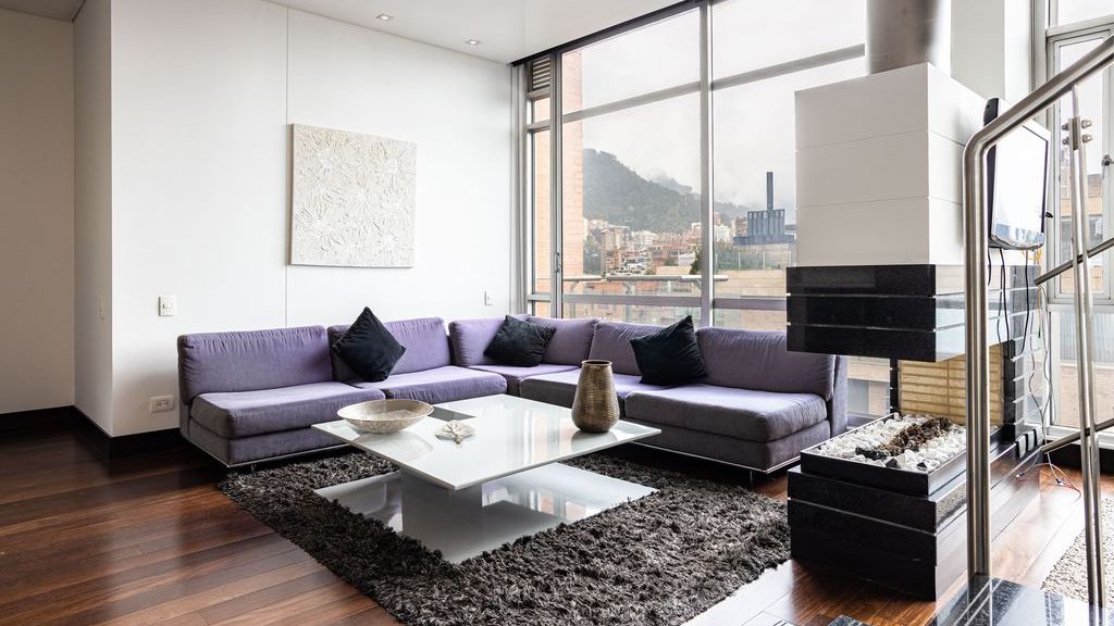 Modern living room with a purple sectional sofa placed in front of floor-to-ceiling windows, a white coffee table, dark rug, and city views.