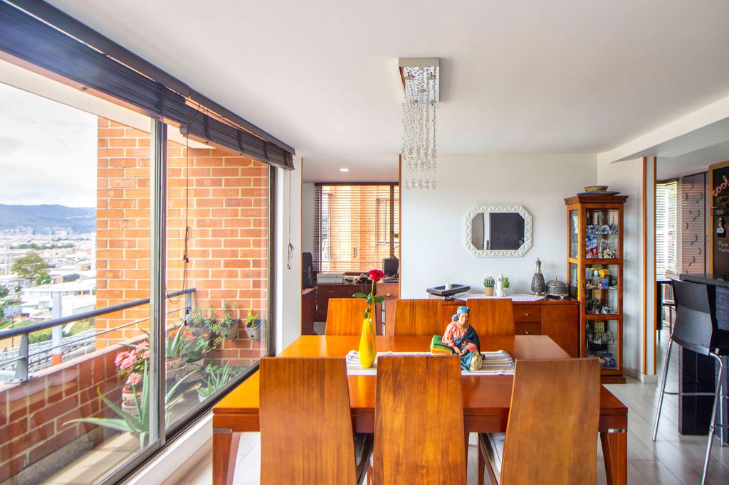 Eclectic dining room with warm wood table, matching chairs, vintage display cabinet, personal decor, balcony plants, and bright natural light.