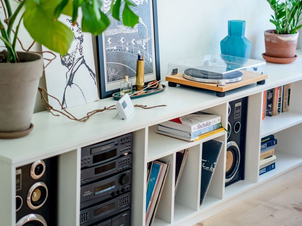 White open media console with stereo equipment, vinyl records, books, plants, framed art, and a record player in a bright living room.