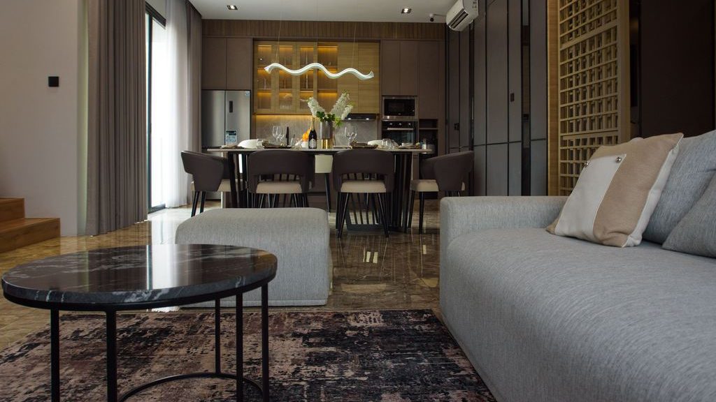 Modern open-plan living and dining room with a gray sofa, round black marble coffee table, patterned rug, dark wood cabinetry, and sculptural pendant lighting.
