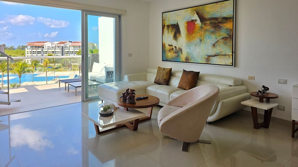 Bright modern living room with a white sectional sofa, curved accent chair, wood coffee tables, and large sliding doors opening to a poolside balcony.