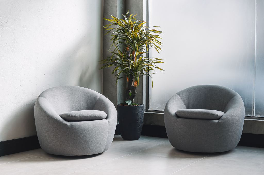 Two gray swivel barrel chairs beside a tall potted plant in a bright modern corner with floor-to-ceiling windows.