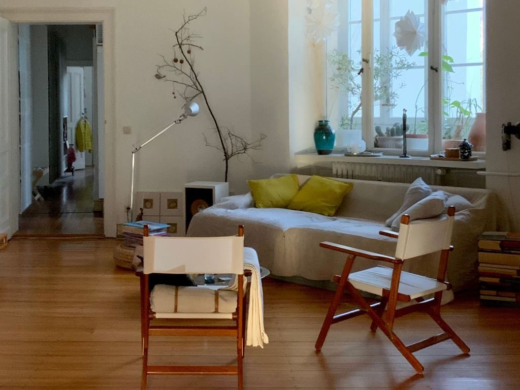 Warm minimalist living room with wood floors, white lounge seating, folding chairs, large windows, plants, and soft natural light.