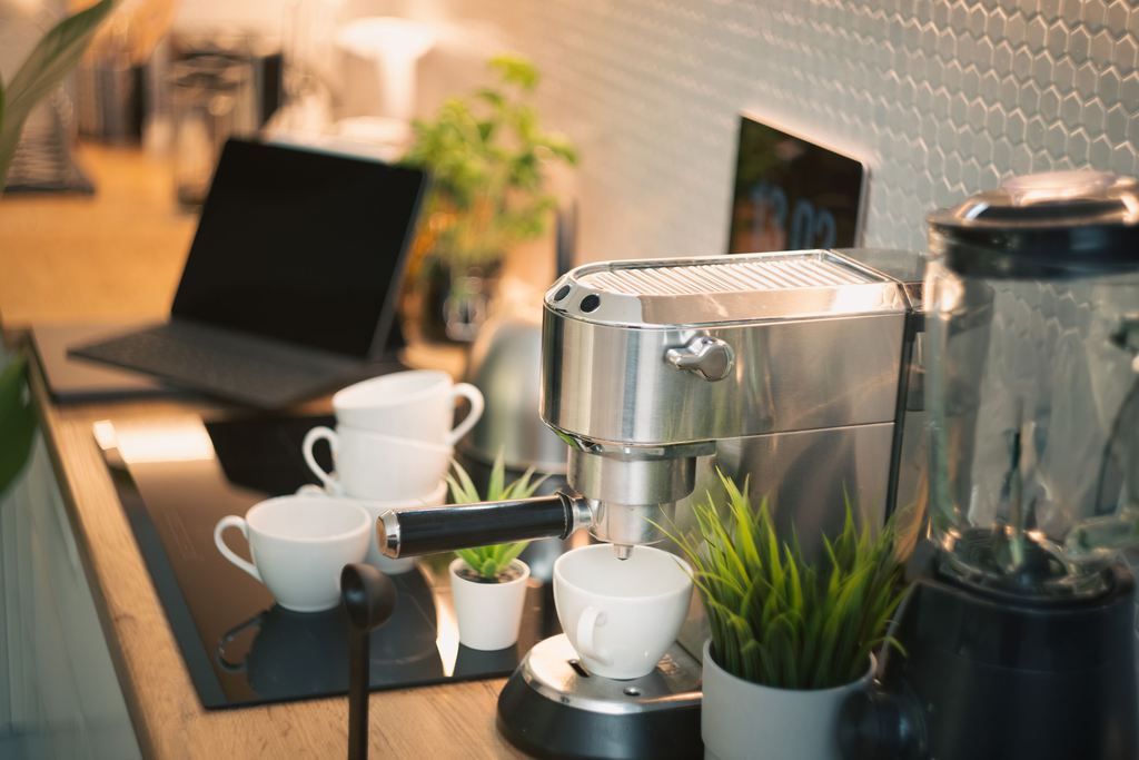 Modern home coffee bar with espresso machine, white cups, blender, plants, and laptop on a warm kitchen countertop.