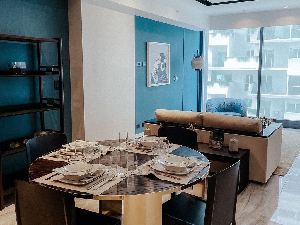 Open-plan rental apartment with round dining table, black chairs, blue accent wall, shelving, and living area near large windows.