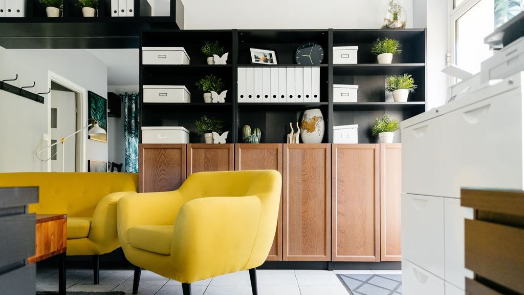 Small home office and living area with black open shelving, wood storage cabinets, white file drawers, and yellow armchairs.
