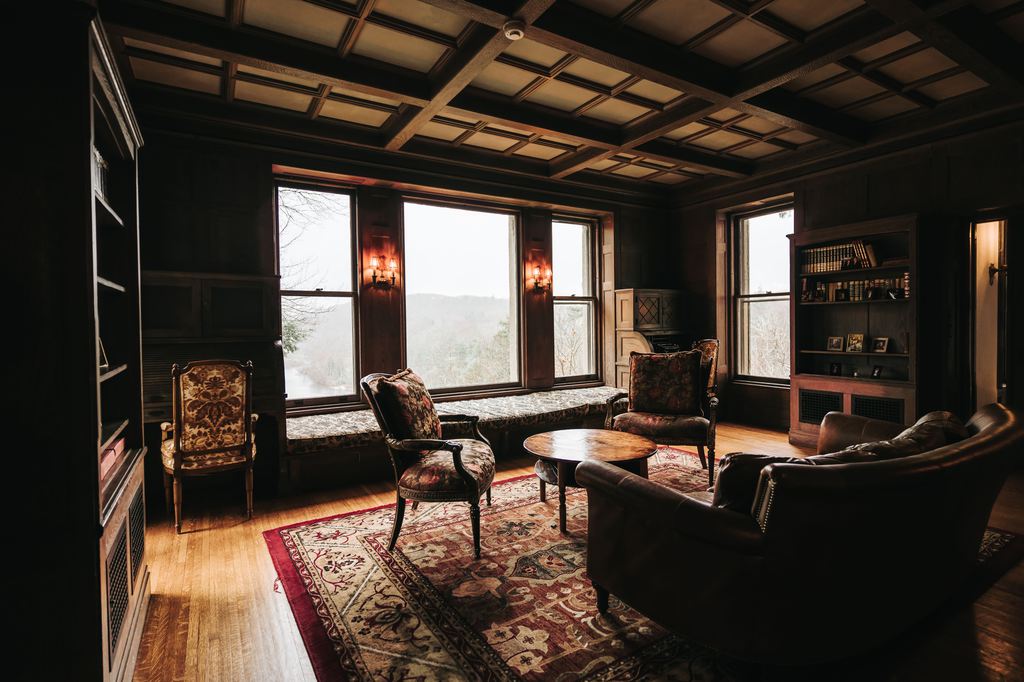 Vintage-style sitting room with dark wood paneling, leather sofa, patterned chairs, round coffee table, red area rug, built-in shelves, and large windows.