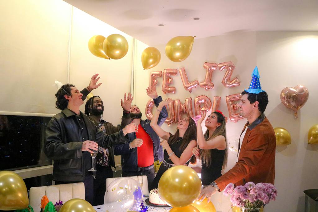 Friends celebrate indoors with gold balloons, party hats, a cake table, and festive wall decorations in a bright living room party setup.