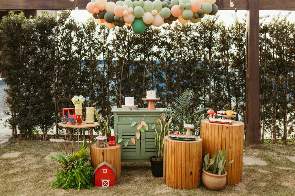 Outdoor party setup with pastel balloon garland, wooden display tables, green cabinet, plants, and small dessert decorations under a covered patio.