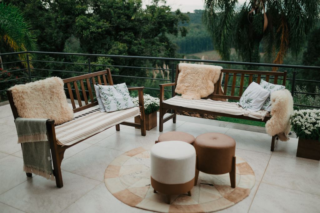 Outdoor patio seating area with wooden benches, round rug, upholstered stools, throw pillows, faux fur accents, and green landscape views.