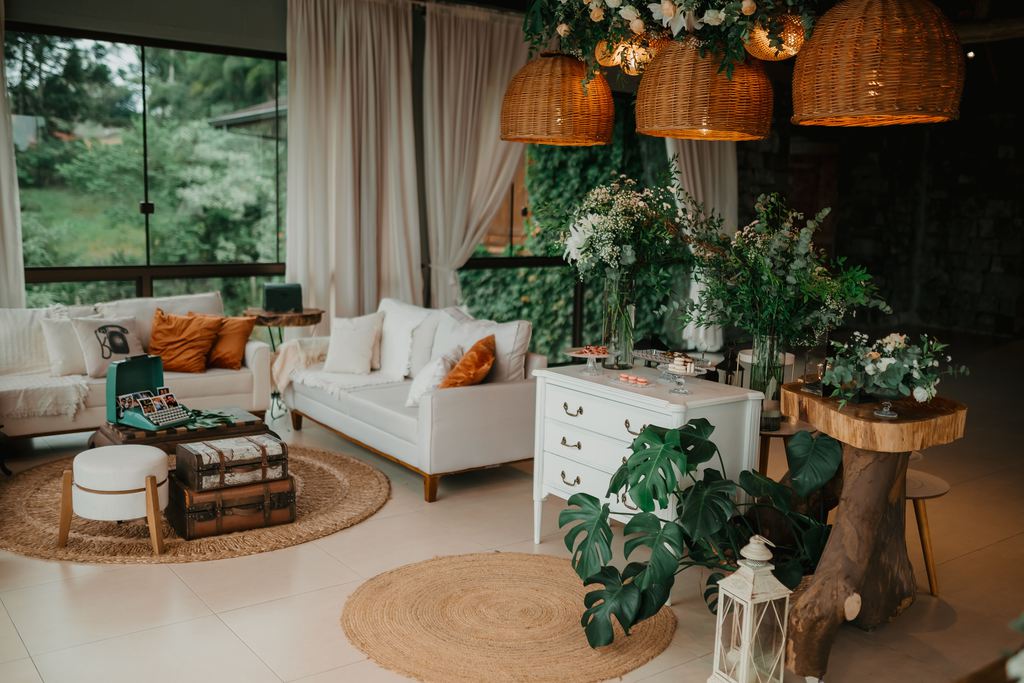 Boho living room with white sofas, woven pendant lights, round jute rugs, leafy plants, wood accents, and warm natural textures near large windows.