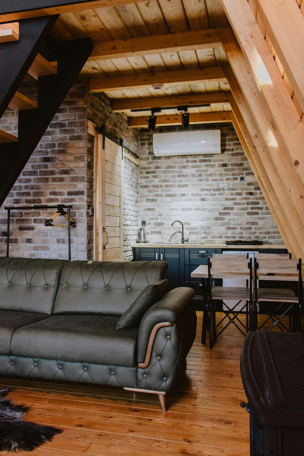 Modern rustic living room with dark leather sofa, exposed brick wall, wood ceiling beams, and compact dining area under warm natural wood tones.