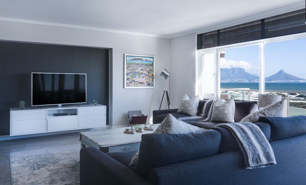 Coastal living room with dark sectional sofa, white media console, light coffee table, and large windows overlooking the ocean.
