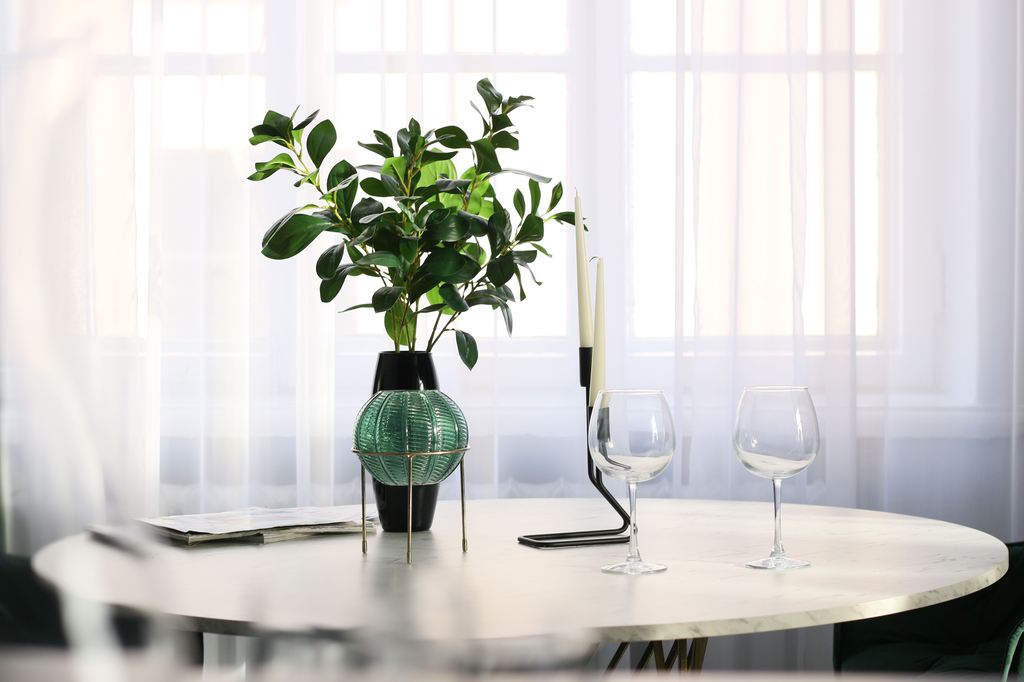 Round white marble dining table with a leafy green centerpiece, two wine glasses, and black candleholders in front of sheer window curtains.