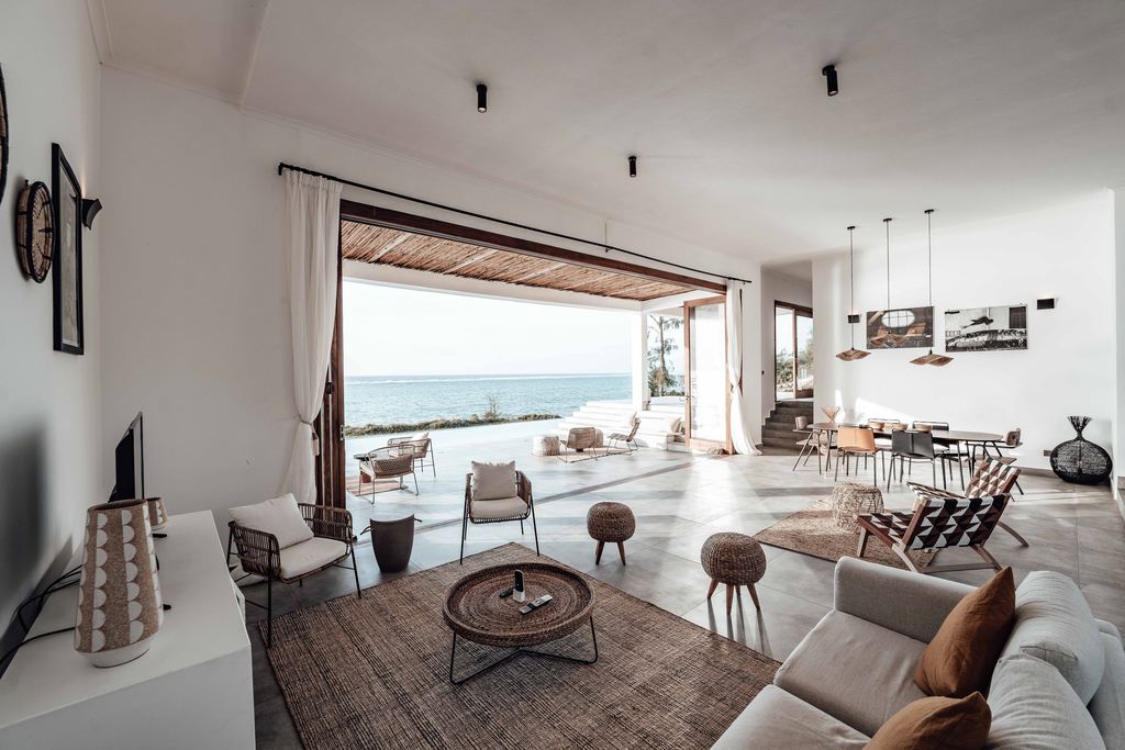 Open coastal living room with neutral sofa, woven furniture, textured rug, dining area, and large sliding doors overlooking the ocean.
