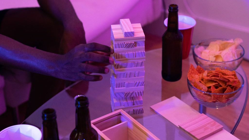Guests play a wooden block-stacking game beside beer bottles, red cups, and bowls of chips on a glass coffee table under purple lighting.