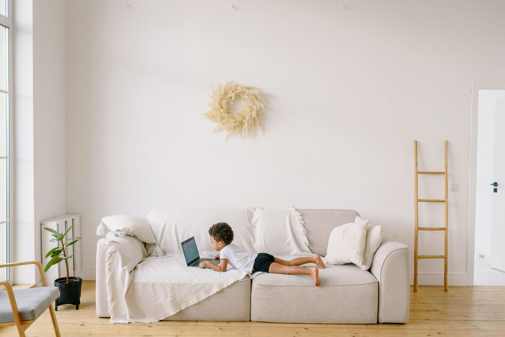 Child lying on a light-colored fabric sofa while using a laptop in a bright living room.