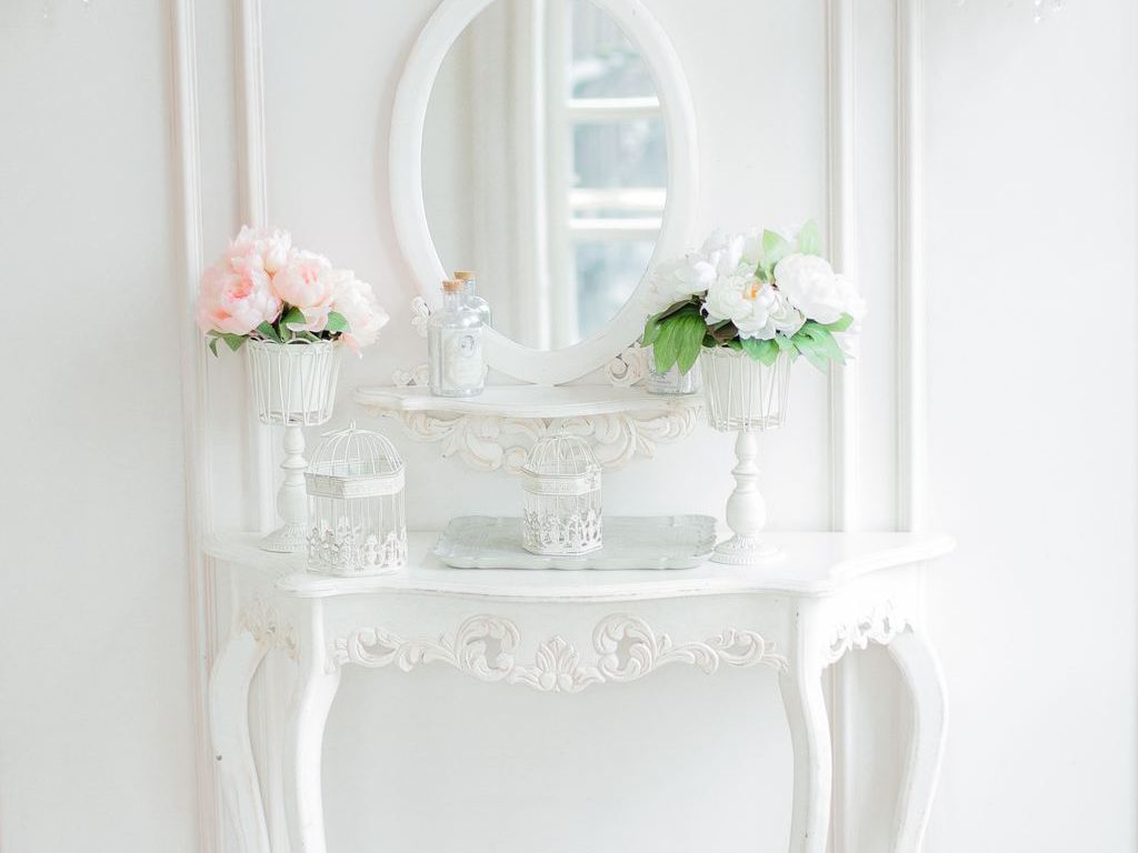 White ornate console table with an oval mirror, floral arrangements, and decorative birdcages in a classic entryway.