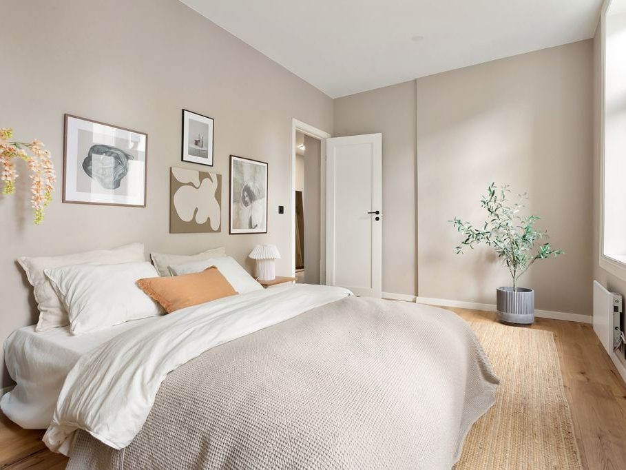 Scandinavian-style bedroom with beige walls, white bedding, light wood flooring, woven rug, framed wall art, and simple natural decor.