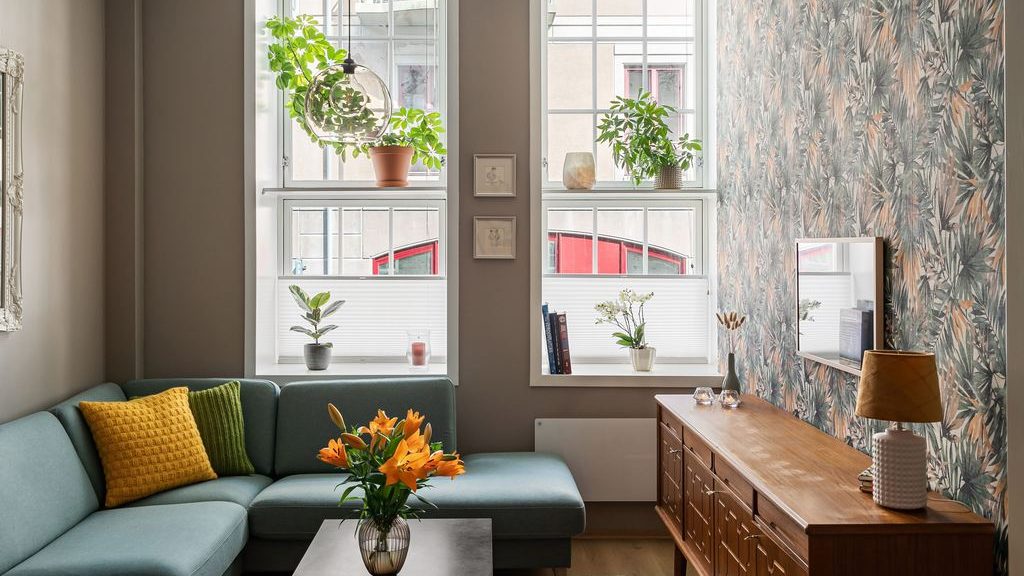 Modern eclectic living room with teal sofa, vintage wood media console, botanical wallpaper, colorful pillows, plants, and layered natural textures.