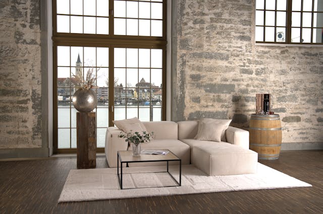 Minimal lake home living room with soft neutral seating, stone-textured walls, wood flooring, and large windows overlooking calm water.