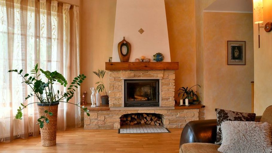 Warm living room with a stone fireplace, wood mantel decor, indoor plants, sheer curtains, and cozy neutral seating.