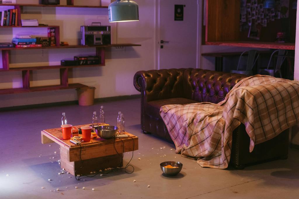 Messy living room after a watch party with popcorn crumbs, cups, bottles, and a blanket draped over a brown leather sofa.