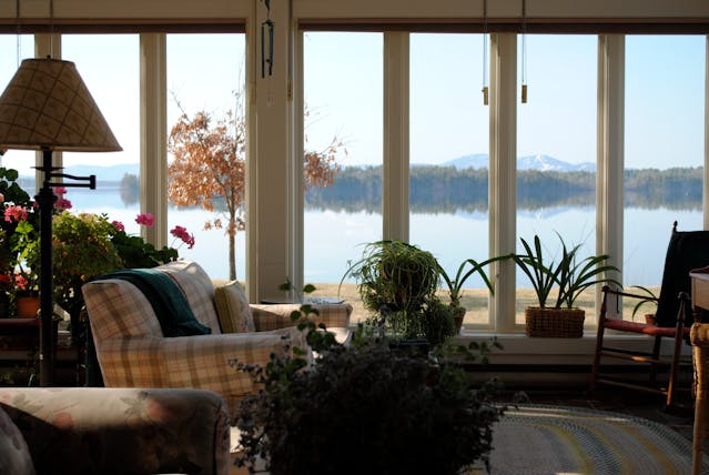 Cozy lake home sitting area with indoor plants, soft lamp lighting, and wide windows framing a peaceful water view.