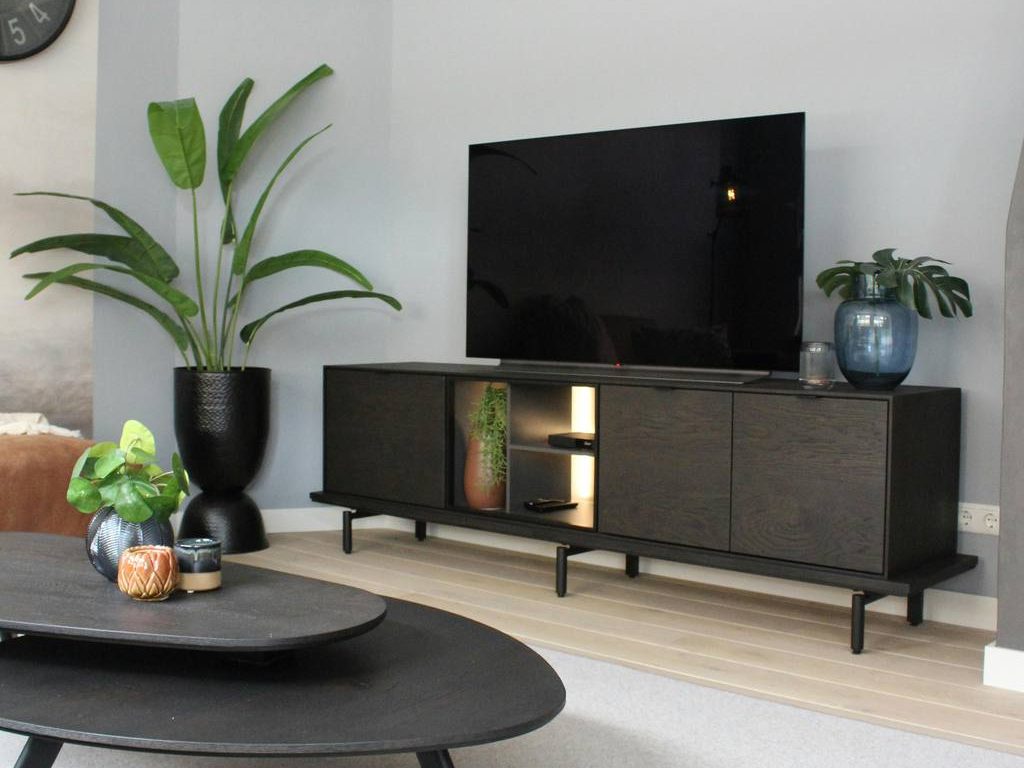 Modern black TV stand with closed storage, open shelf lighting, flat-screen TV, indoor plants, and black coffee table in a calm living room.