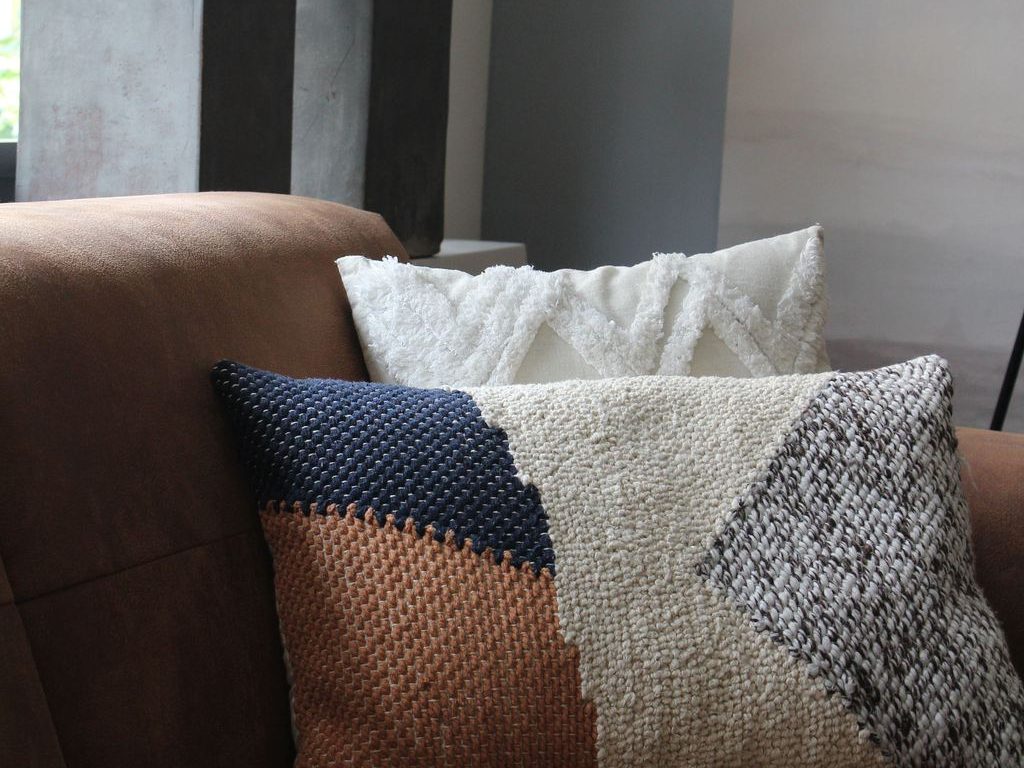 Textured throw pillows on a brown sofa beside a window, with leafy indoor plants adding a cozy living room accent.